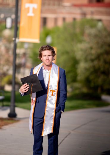 Graduate in blue suit holding a cap on campus.