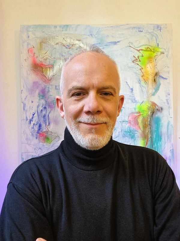 Smiling man with white beard in black turtleneck, standing in front of colorful abstract painting.