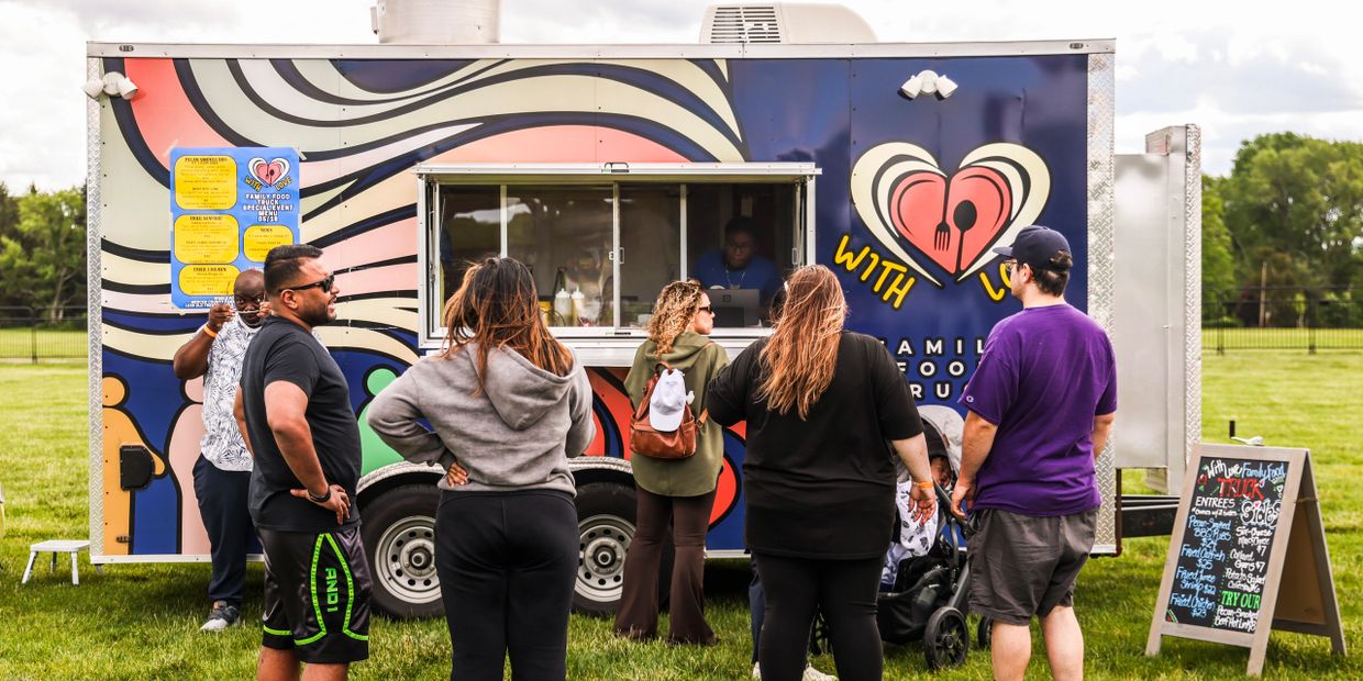 People wait in line at a colorful food truck in a park.