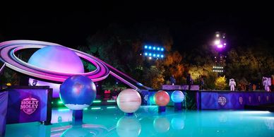 Colorful illuminated planets and Saturn model around a glowing pool at night.