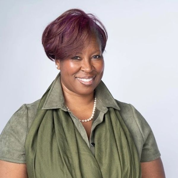 Smiling woman wearing a green outfit and pearl necklace against a plain background.