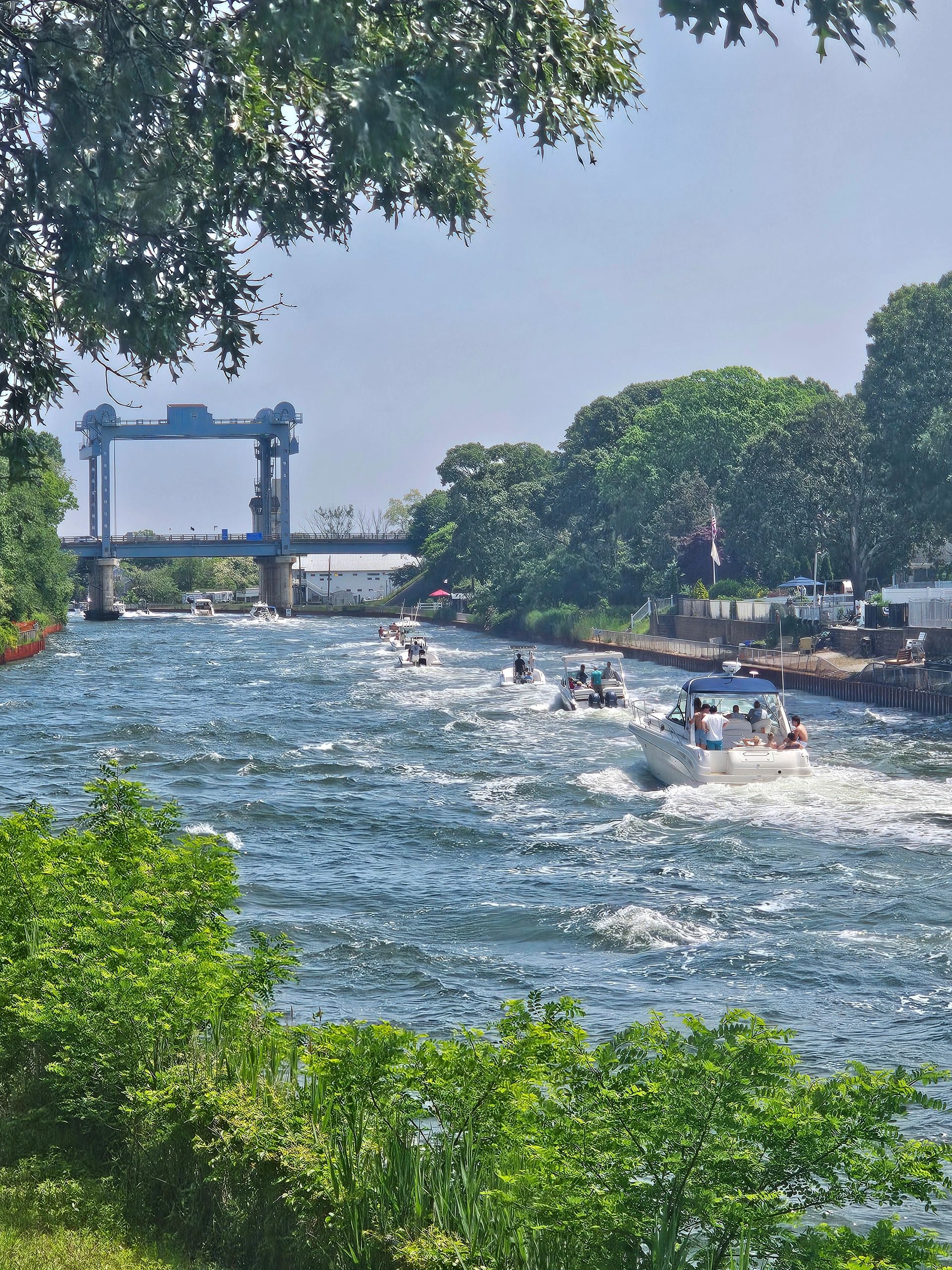 Boats cruising along a river under a blue bridge on a sunny day.