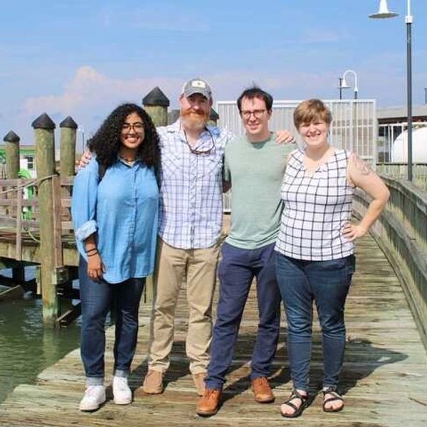 Four friends standing on a wooden dock near water.