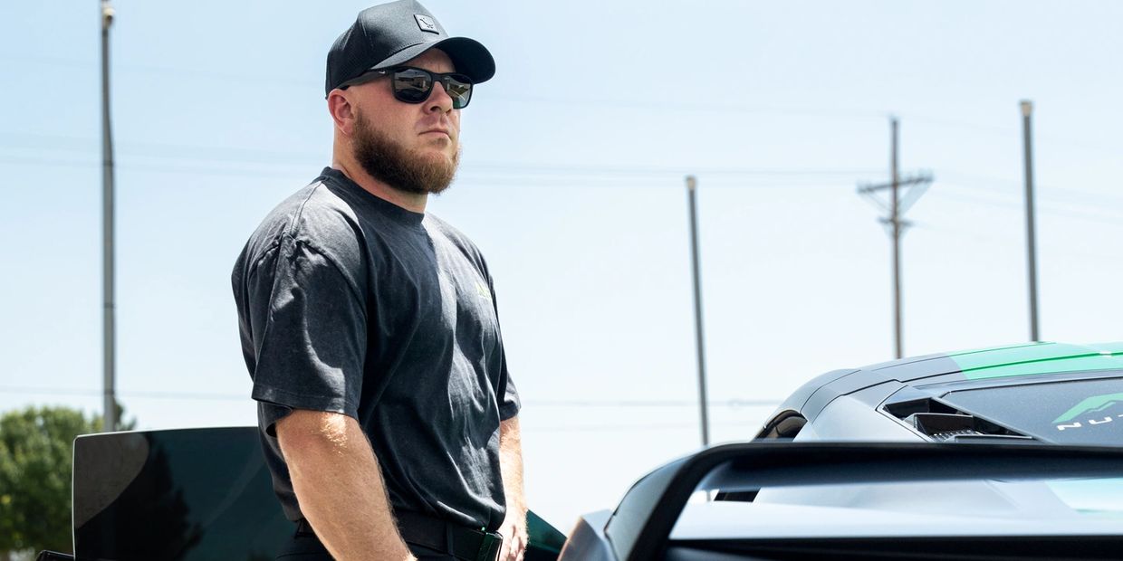 Man in black cap and sunglasses standing beside a sports car under a clear sky.