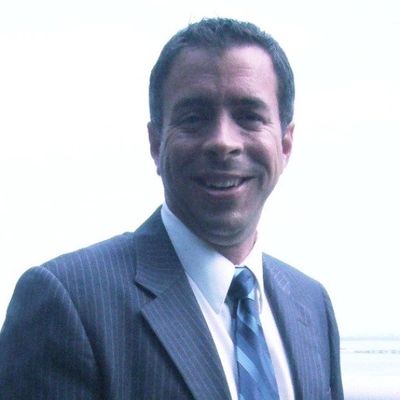 Man in a suit smiling outdoors with a light sky background.