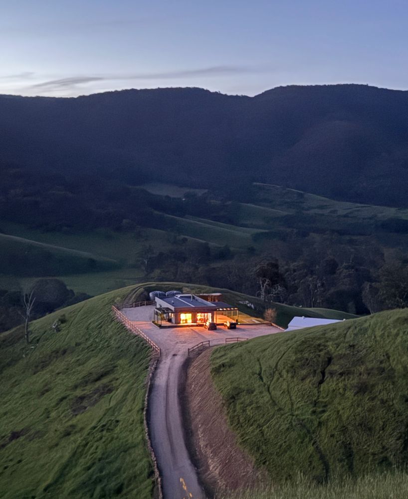 A modern house with glowing lights nestled on a green hill at dusk.
