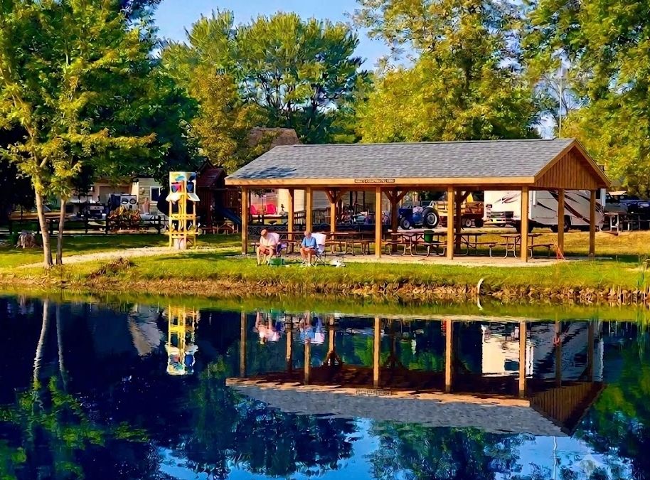 A peaceful pavilion by a lake with two people sitting and trees around.