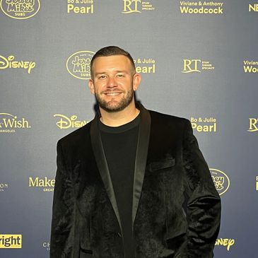 Man in a black velvet jacket posing at an event with branded backdrop.