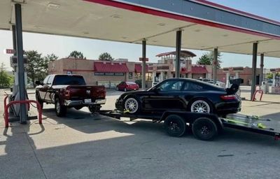 A black sports car is being towed on a trailer by a pickup truck at a gas station.