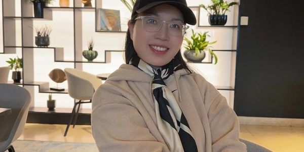 A smiling woman wearing a beige coat, glasses, and a black cap indoors.