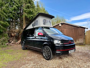 Black camper van with pop-up roof parked by wooden shed and trees.