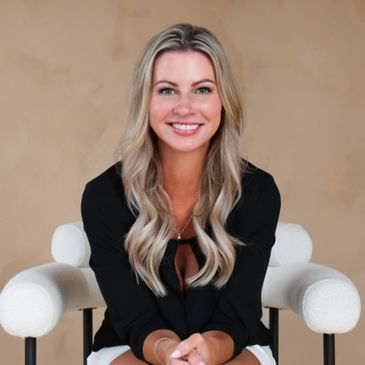 Smiling woman with blonde hair in black top sitting on a white chair.