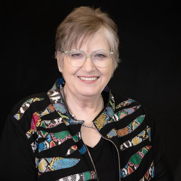 Smiling older woman wearing glasses and a colorful patterned jacket against a black background.