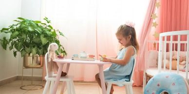A young girl in a blue dress sits at a small table playing with toys in a softly lit room.