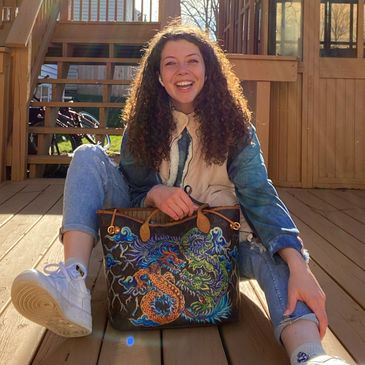 Young woman with curly hair smiling, sitting on a wooden deck holding a colorful dragon-printed bag.