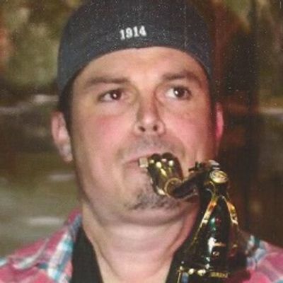 Man playing a saxophone wearing a cap and plaid shirt.