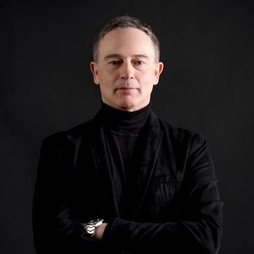 Man in black outfit with arms crossed against black background.