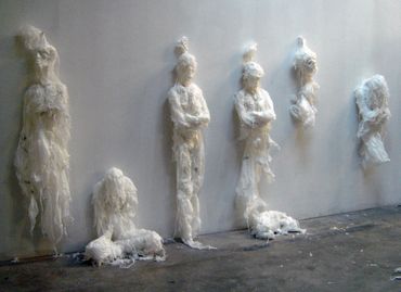 Six humanoid sculptures against a wall made from white trash bags, in varied suspended poses.