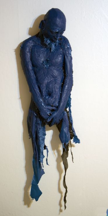 Blue humanoid torso sculpture suspended against a wall with head bowed and hands clasped in front.