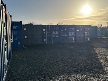 Shipping containers lined up under a bright sun on a gravel ground.