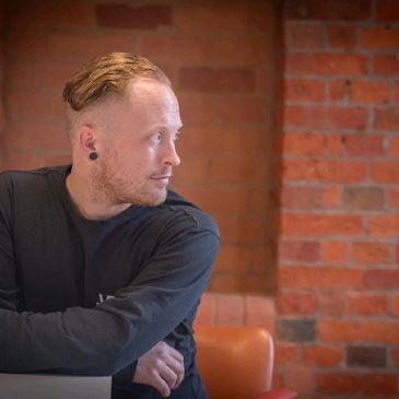 Man with styled hair and earrings looking to the side against a brick wall.
