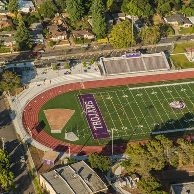 Aerial view of a Trojan football field and track surrounded by trees and houses.