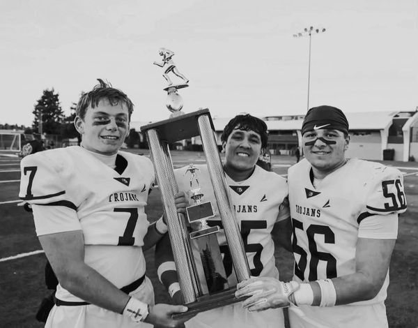 Three football players in Trojans uniforms holding a large trophy on a field.