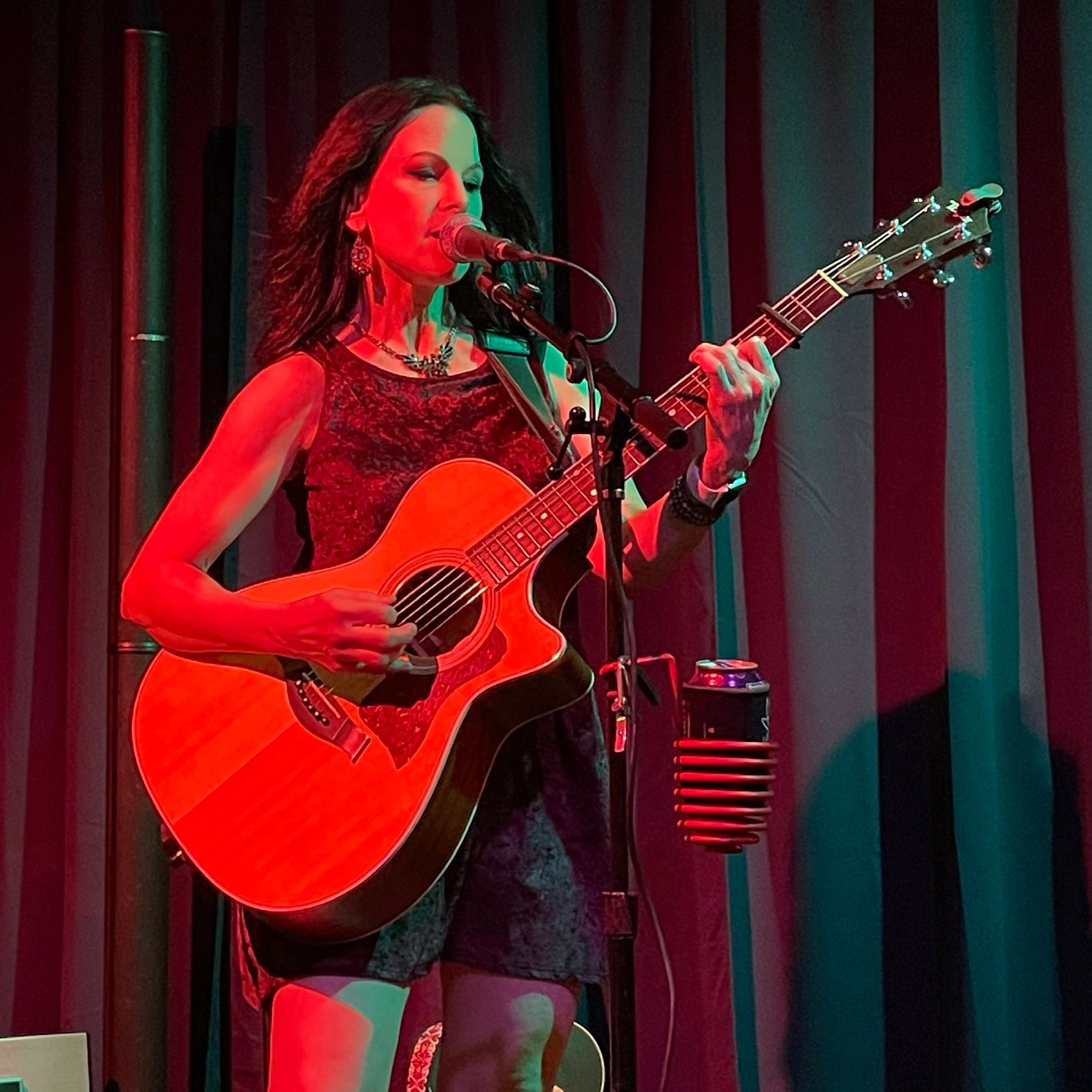 Woman playing acoustic guitar and singing on stage under red and green lights.