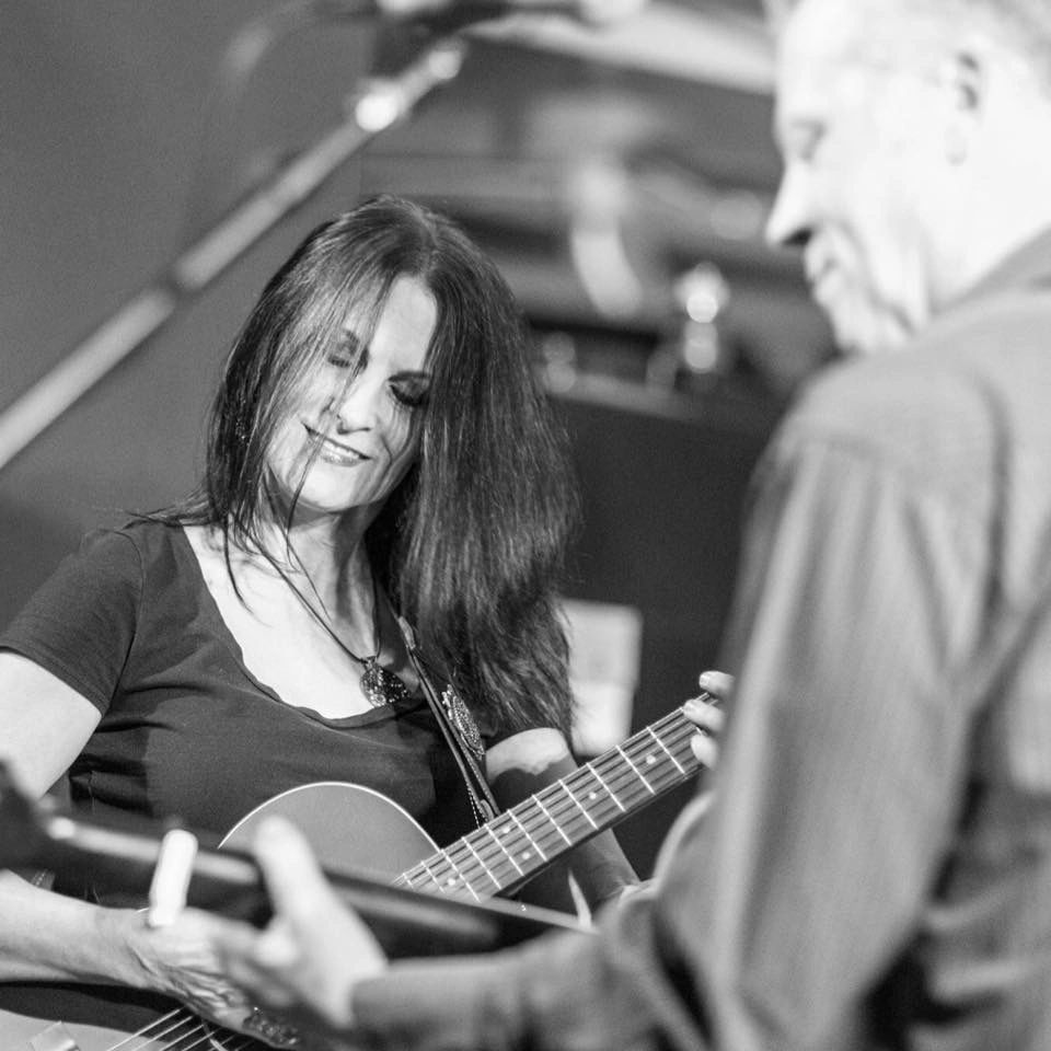 A woman smiling while playing guitar with a man in the foreground.