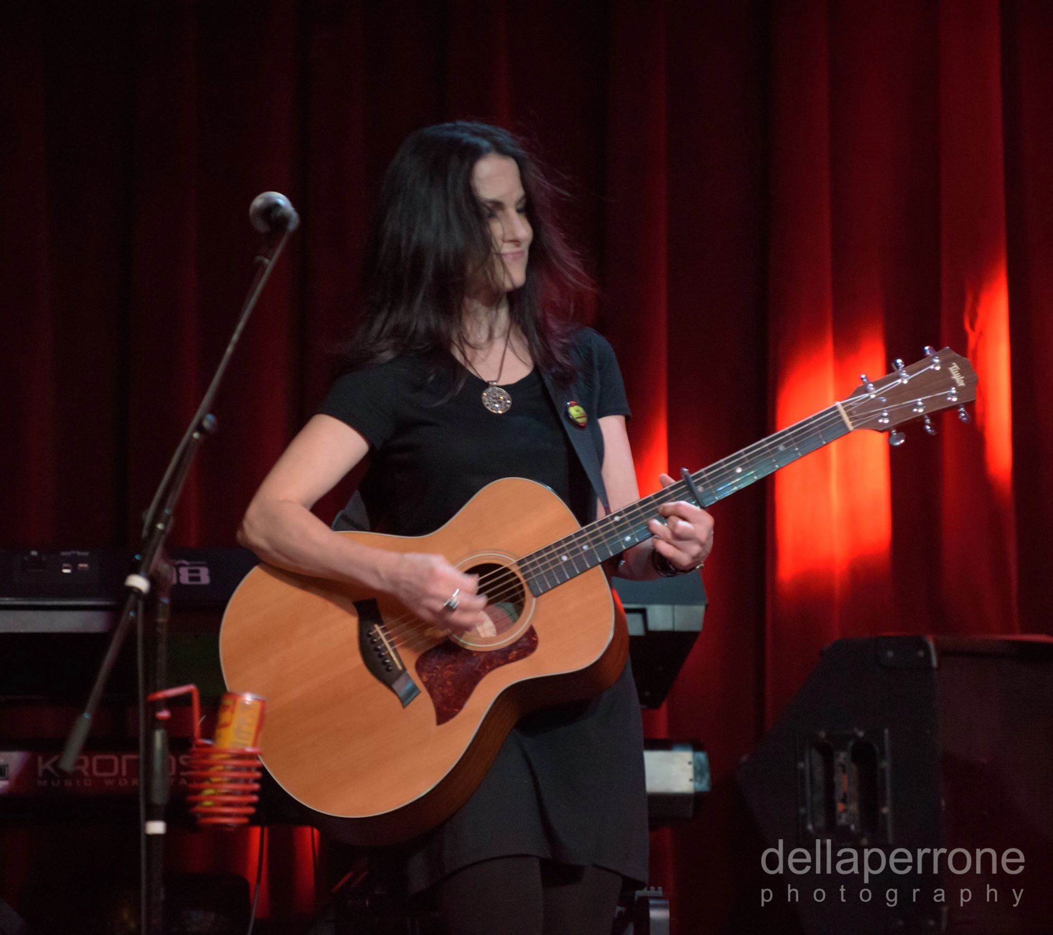 Woman playing acoustic guitar on stage with red curtains in background.