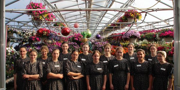 Group of young women in dark dresses standing in a greenhouse filled with flowers.