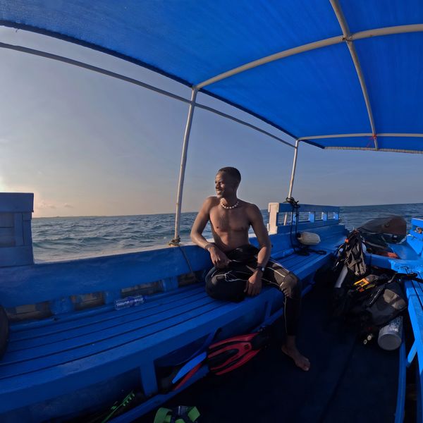 Man sitting on a blue boat under a blue canopy near the ocean.