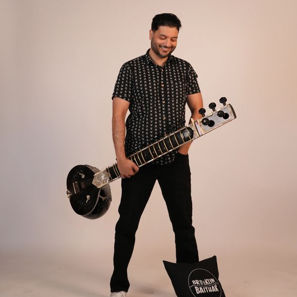 A smiling man holding a sitar against a plain background.