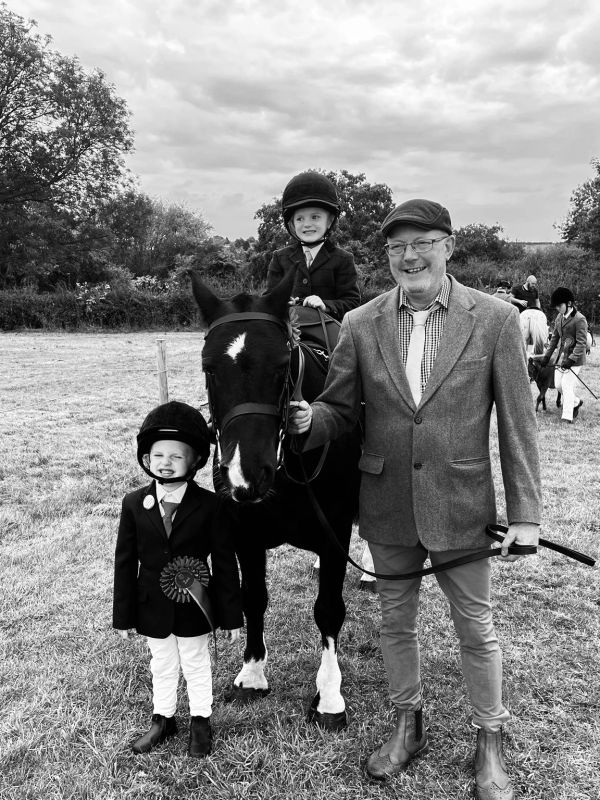 A smiling man and two children in riding attire with a horse outdoors.
