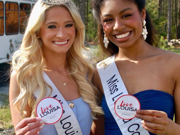 Two smiling women wearing sashes and holding 'Love Louisa' signs outdoors.