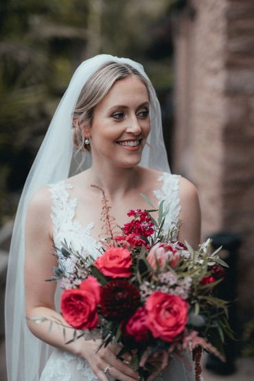 Smiling bride holding vibrant bouquet with red and pink flowers.