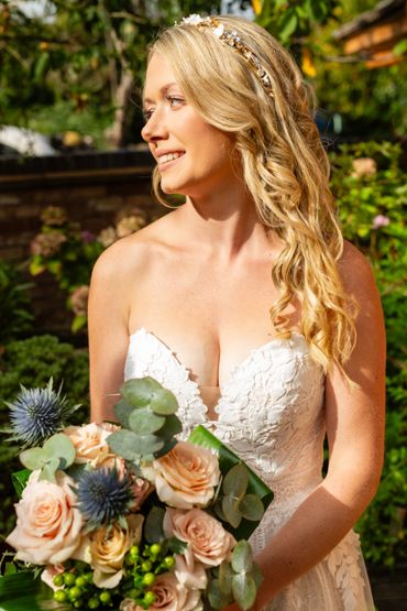 Bride in a white lace dress holding a bouquet, smiling outdoors.