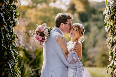 Newlyweds share a joyful embrace and kiss outdoors surrounded by greenery.