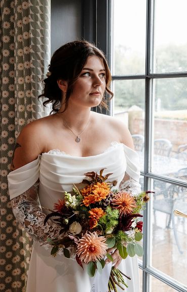 Bride in a white gown holding a vibrant autumn bouquet by a window.