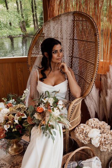 Bride in a white dress holding a bouquet, sitting on a wicker chair.