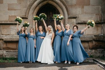 Bride and bridesmaids in blue dresses celebrating with bouquets outside a stone building.