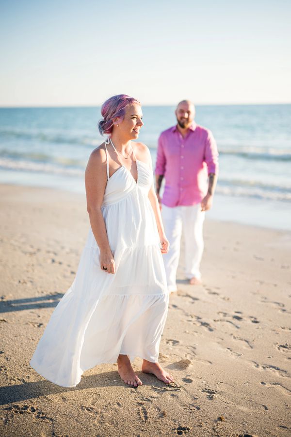 Couple enjoying a sunny day barefoot on the beach, with the woman in a white dress.