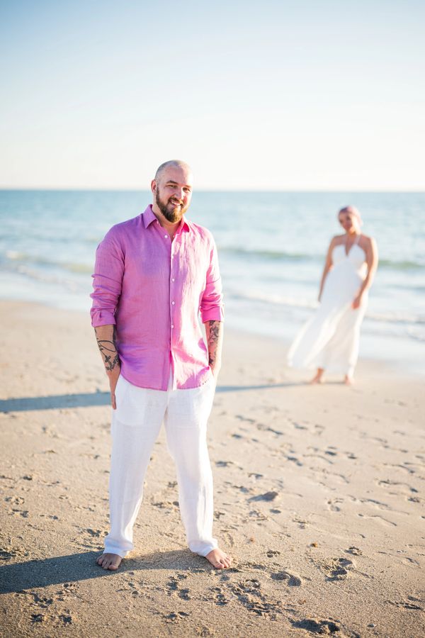 Man in pink shirt and white pants smiling on the beach with a woman in white dress in the background.