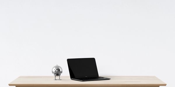 A sleek laptop and a small metallic robot on a wooden desk against a plain white background.