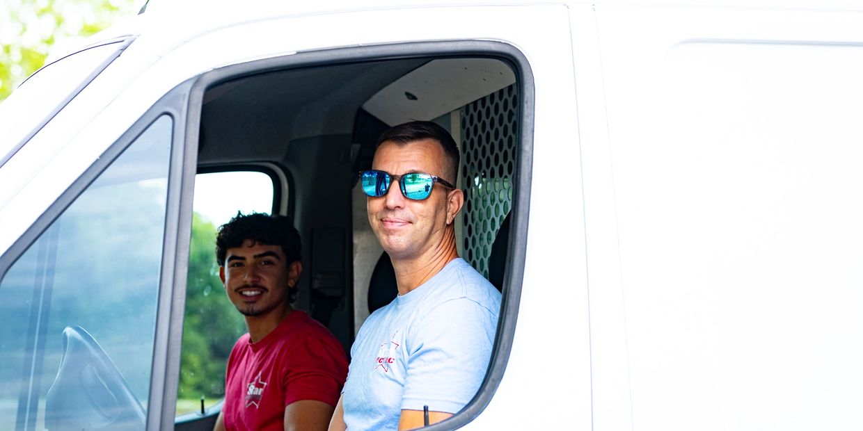 Two men smiling inside a white van, one wearing sunglasses.