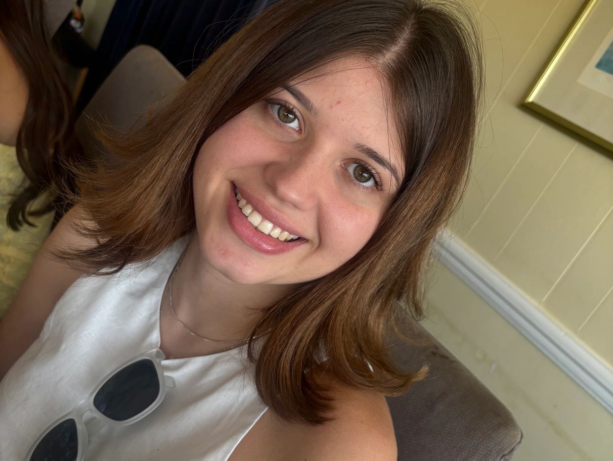 Young woman smiling indoors wearing a white top with sunglasses.