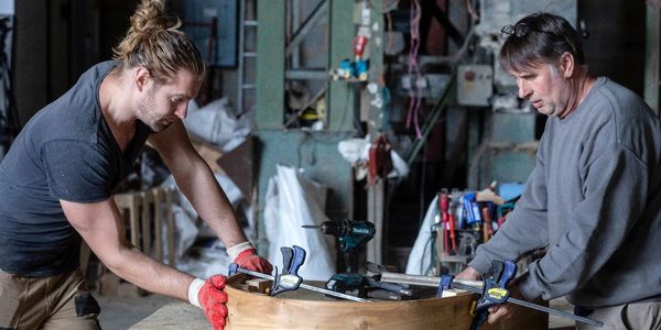 Two men working with wood clamps on a circular wooden piece in a workshop.