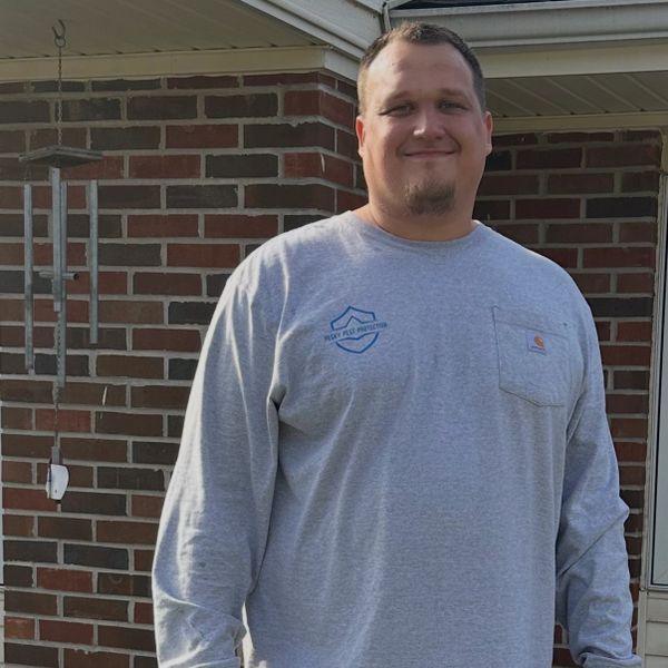 Man smiling in a gray long-sleeve shirt standing outside a brick house.