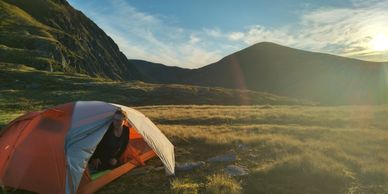 Person emerging from a tent during sunset in a mountainous area.
