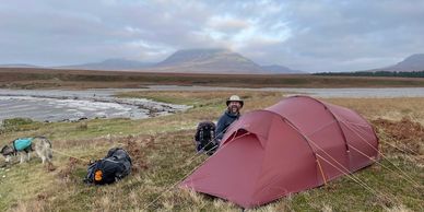 Man camping with a dog beside a red tent on a grassy plain near water.